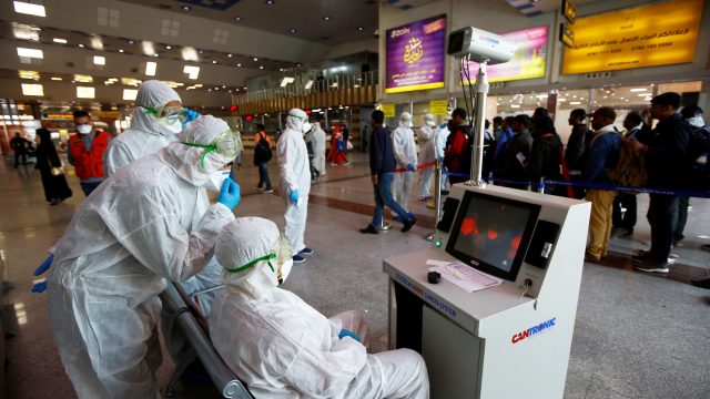 Medical staff in protective gear look at a screen while checking temperatures of passengers